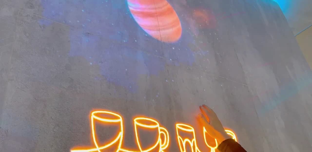 A hand resting on an Econiclay wall where illuminated line drawings of cups and glasses glow from within the stone-look surface, with a planet image above.